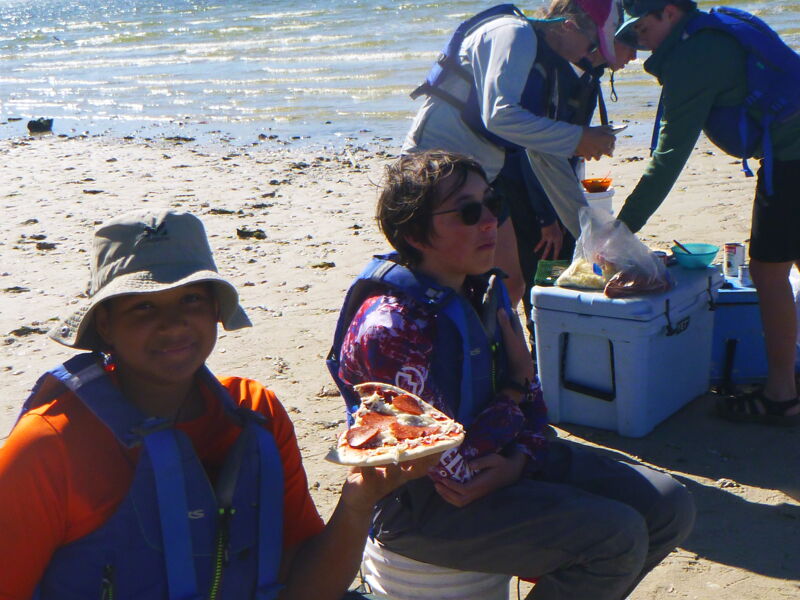 The image shows a group of people on a beach. In the foreground, a person wearing a hat and an orange shirt is holding a slice of pizza. Behind them, another person is sitting and wearing sunglasses and a life vest. In the background, other people are standing near a cooler, possibly preparing food. The scene suggests a casual beach outing with food and relaxation.
